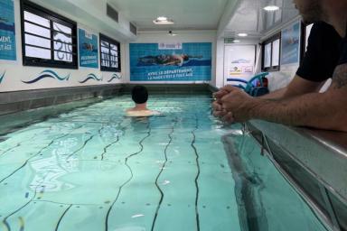 Vue rapprochée du bassin intérieur : un adulte appuyé sur le rebord regarde un enfant dans l’eau. Le bassin est long, avec lignes au fond, affiches bleues sur les murs, lumière artificielle, ambiance calme et éducative.