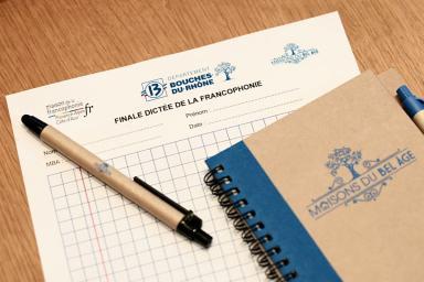 Sur une table en bois reposent une feuille de dictée officielle du Département, un cahier bleu et kraft, ainsi qu’un stylo noir et beige. Le document comporte des espaces pour nom, prénom et date. L’ensemble évoque une préparation à un concours d’écriture et dans le cas présent, une dictée.