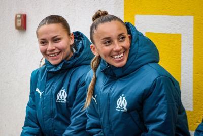 Deux joueuses de l'Olympique de Marseille, souriantes, portant des vestes bleues à l'effigie du club, assises devant un mur blanc décoré de motifs jaunes - Agrandir l'image 12 sur 16, fenêtre modale