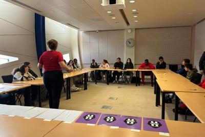 Une salle de réunion avec murs clairs et horloge murale. Une personne debout en rouge parle à un groupe d’environ quinze personnes assises autour de tables disposées en U. Sur les tables, feuilles blanches et affiches violettes avec logos. Un vidéoprojecteur est suspendu au plafond. Les participants semblent attentifs, certains écrivent ou regardent la personne qui parle. - Agrandir l'image 1 sur 5, fenêtre modale