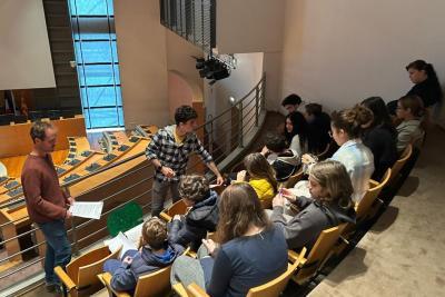 Salle en gradins avec sièges en bois. Une dizaine de personnes assises, certaines écrivent sur des feuilles. Deux personnes debout, l’une tient des documents, l’autre distribue des papiers. En arrière-plan, pupitres et écrans d’une salle de conférence. Grande baie vitrée laissant entrer la lumière. - Agrandir l'image 3 sur 5, fenêtre modale