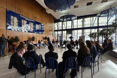 Espace vaste et lumineux avec sol clair et plafond bleu. Une trentaine de personnes assises en cercle sur des chaises bleues. Deux arbres décoratifs et un grand panneau bleu avec noms sur le mur en bois. Une personne au centre semble parler, les autres écoutent. Derrière se trouvent de grandes baies vitrées. - Agrandir l'image 4 sur 5, fenêtre modale