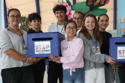 Groupe de sept personnes debout dans un hall lumineux. Elles tiennent deux boîtes bleues avec inscription « STOP Harcèlement ». Derrière, panneaux d’information dont un sur la lutte contre les violences avec une échelle colorée sur le harcèlement et un numéro d’appel. - Agrandir l'image 5 sur 5, fenêtre modale