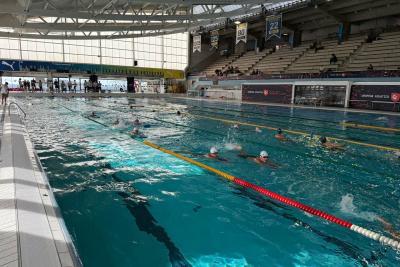 Grand bassin intérieur avec lignes de nage colorées. Plusieurs nageurs équipés de bonnets et lunettes nagent dans des couloirs. Gradins visibles en arrière-plan avec quelques spectateurs. Structure moderne avec toit métallique et larges baies vitrées. - Agrandir l'image 4 sur 5, fenêtre modale