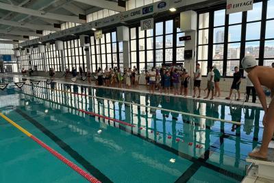 Piscine intérieure avec couloirs de nage. Une personne en bonnet se tient prête à plonger sur un plot au premier plan. En arrière, groupe aligné le long du bassin, semblant attendre leur tour. Grandes fenêtres laissant entrer la lumière, ambiance de compétition. - Agrandir l'image 5 sur 5, fenêtre modale