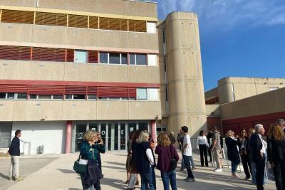 Un groupe de personnes se tient devant un bâtiment à plusieurs étages, identifiable par des éléments rouges sur la façade. Il s’agit probablement d’une visite officielle. - Agrandir l'image 4 sur 6, fenêtre modale