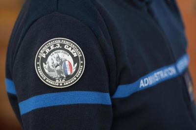 Un adulte en tenue bleu foncé est photographié de côté. Sur sa manche, un écusson rond indique un service pénitentiaire avec des symboles officiels. Une bande bleue court sur la veste. L’environnement intérieur est lumineux, probablement un hall où circulent d’autres agents. - Agrandir l'image 7 sur 18, fenêtre modale