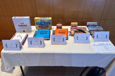 Une table recouverte d’une nappe blanche présente divers lots : jeux de société, livres, coffrets et boîtes colorées. Des étiquettes blanches portant « Les Lauréats » sont alignées devant les récompenses. L’ensemble est bien ordonné, prêt pour une remise de prix. - Agrandir l'image 4 sur 5, fenêtre modale