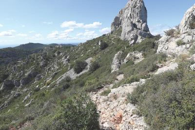 Un sentier rocailleux serpente à travers une zone montagneuse, menant à une formation rocheuse proéminente. Le paysage est parsemé de buissons verts sous un ciel partiellement nuageux. - Agrandir l'image 2 sur 4, fenêtre modale