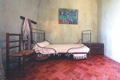 Chambre simple aux murs clairs, avec un lit en fer forgé recouvert de draps blancs, une chaise en bois à assise tressée, quelques vêtements suspendus à des crochets, et un tableau de fleurs accroché au-dessus du lit. Le style évoque une époque ancienne ou une reconstitution historique. - Agrandir l'image 3 sur 6, fenêtre modale