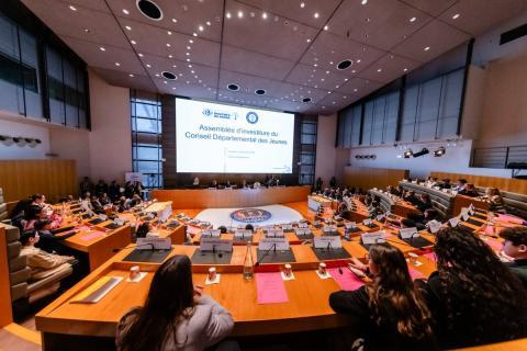 Salle de réunion officielle vue de l’arrière. De nombreux jeunes sont assis autour de pupitres en bois, regardant un grand écran affichant une assemblée d’investiture. Micros, dossiers et bouteilles d’eau visibles.