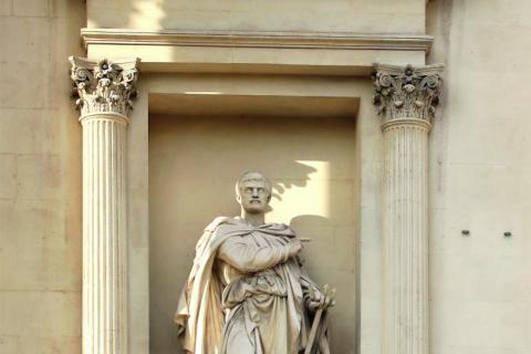 statue d'un homme paré d'une cape par dessus sa tunique et son pantalon, pointant son doigt droit dans une direction tandis que sa main gauche tient un petit trident