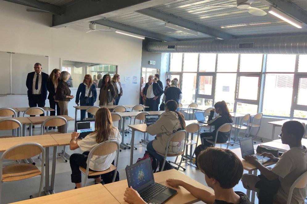 Des élèves sont assis à leur bureau dans une salle de classe, travaillant sur des ordinateurs portables. À l’arrière-plan, un groupe d’adultes observe la scène, probablement en visite pédagogique.